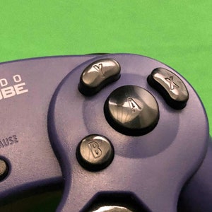 May include: A purple Nintendo GameCube controller with black buttons labeled A, B, X, and Y. The controller has a black start/pause button and the text "Nintendo Cube" is printed on the front.
