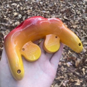 Puede incluir: Un mando de GameCube en degradado rojo, naranja y amarillo. El mando tiene dos botones circulares amarillos con el logotipo de Nintendo y el texto "GameCube Controller". El mando se sostiene en una mano.