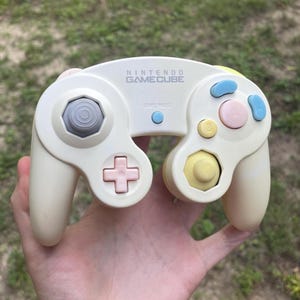 May include: A cream-colored Nintendo GameCube controller featuring a gray analog stick, a pink D-pad, a yellow C-stick, a pink A button, two light blue action buttons, and a small light blue start/pause button centered below the logo text.