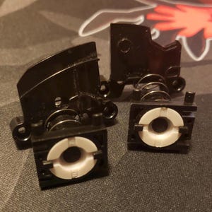 Peut inclure: Quatre composants en plastique noir avec des inserts circulaires blancs et des ressorts métalliques. Les pièces sont disposées sur une surface sombre, avec un arrière-plan flou avec des formes rouges et blanches.