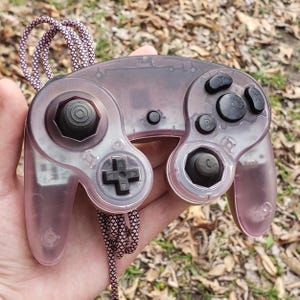 Puede incluir: Un mando de videojuegos rosa transparente con botones negros y un joystick negro. El mando tiene un cable con un patrón rosa y blanco. El mando se sostiene en una mano.