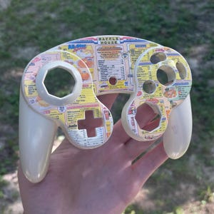 May include: A cream-colored video game controller shell featuring a Waffle House menu graphic printed across the front surface. The shell includes cutouts for buttons and joysticks, displaying text such as All-Star Specials and various menu items.