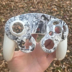 Könnte beinhalten: Weißer GameCube-Controller mit einem Manga-Design in Schwarz und Weiß. Der Controller zeigt Manga-Panels und den Text "THE WORLD REMEMBERS ME AT LONG LAST!!". Er hat ein glänzendes Finish.