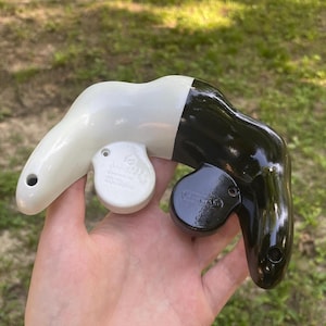 May include: A two-toned video game controller shell featuring a glossy white left side and a glossy black right side. The controller has a distinct ergonomic shape with circular protrusions for the analog sticks and visible screw holes.