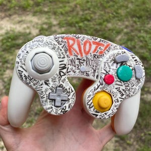Puede incluir: Un mando de videojuegos blanco con un patrón de grafiti negro personalizado con la palabra RIOT repetida por toda la superficie. El mando incluye un gráfico rojo grande de RIOT, un stick C amarillo, un botón A verde y controles direccionales grises.
