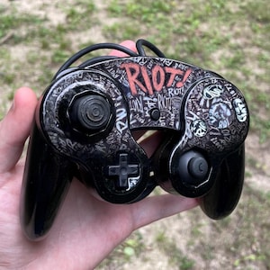May include: A black video game controller featuring a custom graphic wrap with the word RIOT! in red text at the top center. The surface is covered in a repeating pattern of the word RIOT! in white and gray, with black buttons and a black joystick.