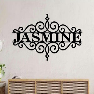 Può includere: Arte da parete in metallo nero con la scritta "JASMINE" in un carattere decorativo. Il nome è centrato all'interno di un ornato disegno a volute. L'opera d'arte è montata su una parete bianca sopra un mobile in legno.