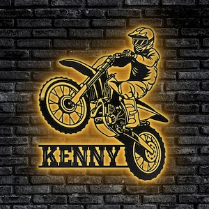 Può includere: Una silhouette in bianco e nero di un pilota di motocross su una moto da cross, con il nome "KENNY" in un font stilizzato sotto. L'immagine è su uno sfondo di muro di mattoni scuri.