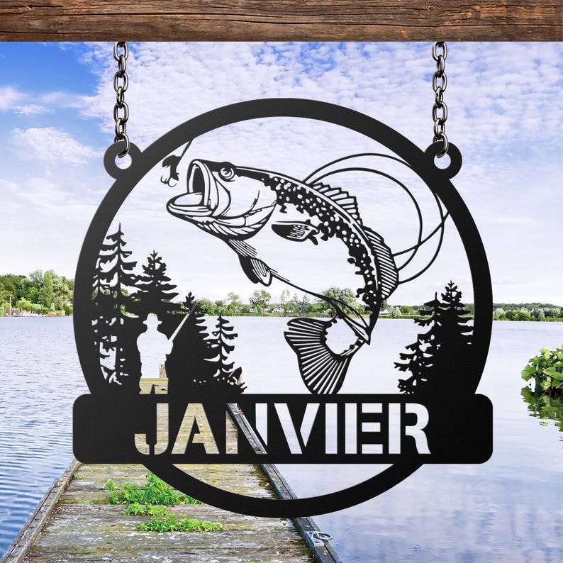 May include: Black metal sign featuring a leaping fish with a hook in its mouth, set within a circular frame. The sign includes a silhouette of a fisherman and the name "JANVIER" in bold letters. The sign hangs from chains against a lake backdrop.