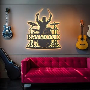 May include: A wall-mounted metal art piece featuring a drummer silhouette with the name "RAYMOND" illuminated in a warm glow. The artwork is surrounded by guitars and a red sofa, creating a music-themed room.