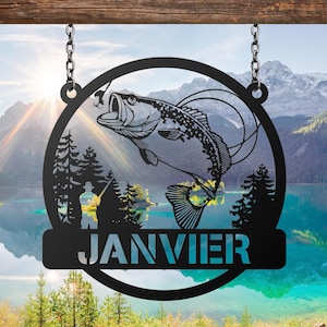 May include: Black metal sign featuring a leaping bass fish design within a circular frame, suspended by chains. The sign includes the name "JANVIER" in a rectangular banner. The background shows a scenic lake and mountain landscape.
