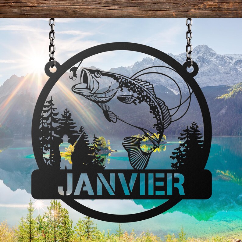 May include: Black metal sign featuring a leaping bass fish design within a circular frame, suspended by chains. The sign includes the name "JANVIER" in a rectangular banner. The background shows a scenic lake and mountain landscape.