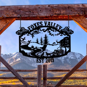 May include: Black metal sign with a rustic design featuring a river, trees, animals, and a tractor. The sign reads "Foxes Valley Est 2019" and is hanging from a wooden gate.