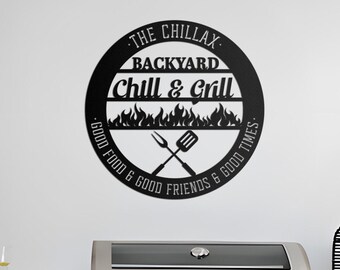 Könnte beinhalten: Schwarzes Metallschild mit den Worten "The Chillax Backyard Chill & Grill" in einem kreisförmigen Design. Das Schild enthält gekreuzte Gabel und Spatel, Flammen und den Text "Good Food & Good Friends & Good Times."
