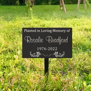 K&ouml;nnte beinhalten: Schwarzes Garten Schild aus Metall mit wei&szlig;em Text, der "Gepflanzt in liebevoller Erinnerung an Rosalie Bradford 1976-2022" lautet. Das Schild hat unten ein dekoratives Blumenmuster.