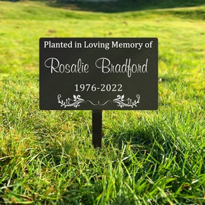 K&ouml;nnte beinhalten: Schwarzes Garten Schild aus Metall mit wei&szlig;em Text, der "Gepflanzt in liebevoller Erinnerung an Rosalie Bradford 1976-2022" lautet. Das Schild hat unten ein dekoratives Blumenmuster.