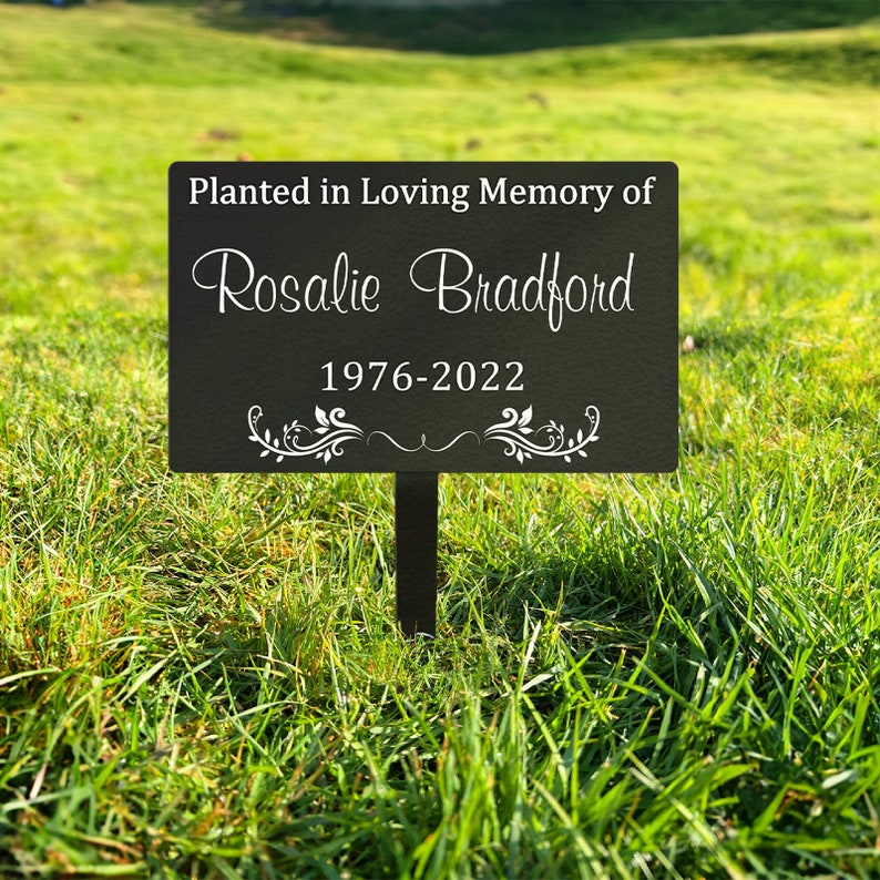 K&ouml;nnte beinhalten: Schwarzes Garten Schild aus Metall mit wei&szlig;em Text, der "Gepflanzt in liebevoller Erinnerung an Rosalie Bradford 1976-2022" lautet. Das Schild hat unten ein dekoratives Blumenmuster.
