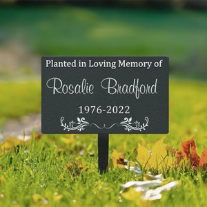 K&ouml;nnte beinhalten: Schwarzes Garten Schild aus Metall mit wei&szlig;em Text, der "Gepflanzt in liebevoller Erinnerung an Rosalie Bradford 1976-2022" lautet. Das Schild hat ein dekoratives Blumenmuster.