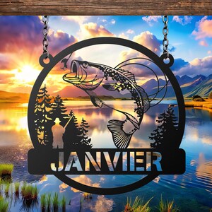 May include: Black metal sign featuring a leaping fish silhouette within a circular frame, suspended by chains. The sign includes a fisherman silhouette, trees, and the word "JANVIER" in a banner, set against a scenic lake and sunset background.
