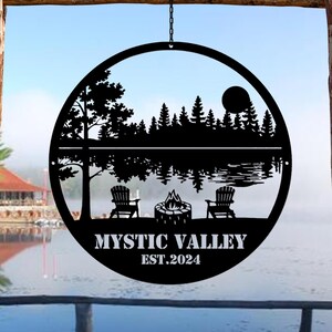 May include: Black metal silhouette of a lake scene with trees, a campfire, and two chairs. The text "Mystic Valley Est. 2024" is below the image.