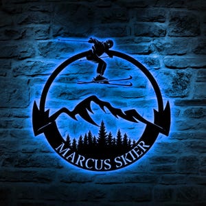 May include: Black metal silhouette of a skier jumping over a mountain range with a forest in the foreground. The silhouette is inside a circle with the text "Marcus Skier" at the bottom.