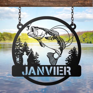 May include: Black metal sign with a circular design featuring a leaping fish, a fisherman, and pine trees. The word "JANVIER" is displayed in white letters on a rectangular bar at the bottom. The sign hangs from a wooden beam with chains.