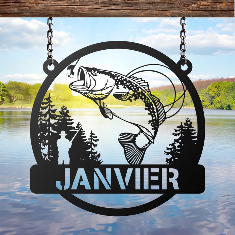 May include: Black metal sign with a circular design featuring a leaping fish, a fisherman, and pine trees. The word "JANVIER" is displayed in white letters on a rectangular bar at the bottom. The sign hangs from a wooden beam with chains.
