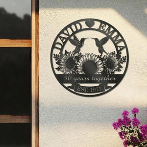 May include: Black metal wall art with the names "David" and "Emma" above two hummingbirds and sunflowers. The text "50 years together" and "EST. 1975" are also included in the design. The art is circular in shape.