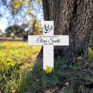 Puede incluir: Una cruz conmemorativa blanca con el texto "FOREVER IN OUR HEARTS" y el nombre "Clara Smith" con los años 1955-2023. Un gráfico de paloma en la parte superior. Una flor amarilla crece en la base de la cruz. La cruz está en la hierba.