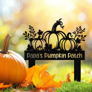 May include: A black metal sign with the text "Papa's Pumpkin Patch" and a decorative design of three pumpkins and foliage. An orange pumpkin and fallen leaves are in the foreground.
