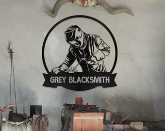 May include: Black and white silhouette of a welder wearing a welding mask and holding a welding torch. The silhouette is inside a circle with the text "GREY BLACKSMITH" on a banner below the circle.