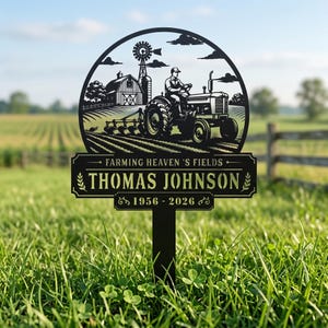 Könnte beinhalten: Schwarzer Metall-Gartenstecker mit einem Traktor, der ein Feld pflügt, einer Scheune und einer Windmühle. Der Text lautet "Farming Heaven's Fields" und "Thomas Johnson 1956-2026". Der Stecker steht in einem grasbewachsenen Feld.