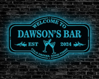 May include: A neon sign with the text "Welcome to Dawson's Bar Est 2024 Cold Drinks, Good Times" and a graphic of two martini glasses. The sign is black with white lettering and a blue neon glow.