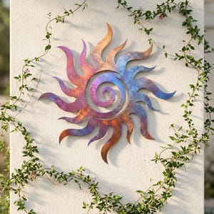 May include: A circular metal wall art piece shaped like a sun with a central spiral. The surface features a gradient of iridescent colors, transitioning from warm copper and orange tones on the left to cool blue and purple hues on the right.