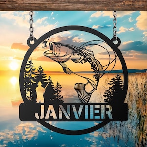 May include: Black metal fishing sign with a circular design featuring a leaping fish, a fisherman, and pine trees against a sunset lake backdrop. The word "JANVIER" is displayed on a lower banner, suspended by chains.
