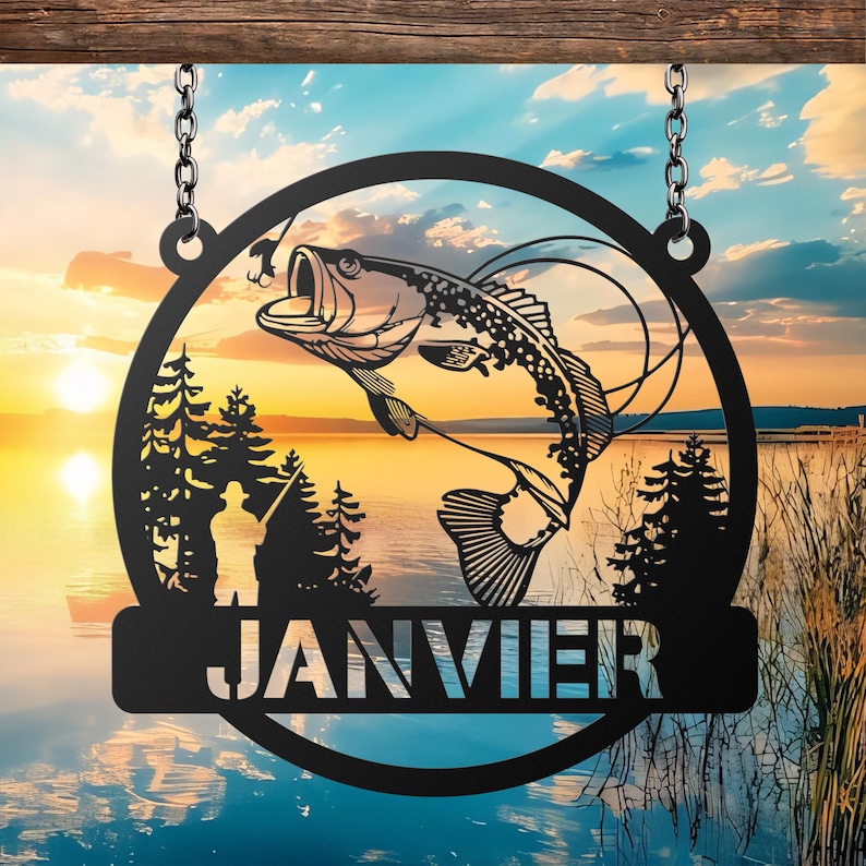 May include: Black metal fishing sign with a circular design featuring a leaping fish, a fisherman, and pine trees against a sunset lake backdrop. The word "JANVIER" is displayed on a lower banner, suspended by chains.