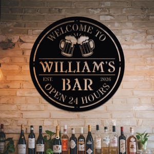Puede incluir: Un letrero de bar de metal negro con el texto "WELCOME TO WILLIAM'S BAR" y "OPEN 24 HOURS". El letrero presenta dos jarras de cerveza y el año de fundación, 2026. Es redondo y está montado en una pared de ladrillo.