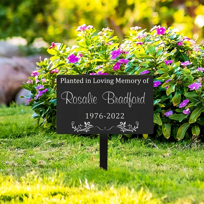 K&ouml;nnte beinhalten: Schwarzes Metall-Gartenschild mit wei&szlig;em Text, der "Gepflanzt in liebevoller Erinnerung an Rosalie Bradford 1976-2022" lautet. Das Schild steht in einem grasbewachsenen Bereich mit rosa Blumen.
