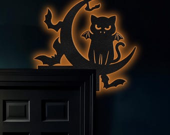 May include: Black metal silhouette of a cat with bat wings sitting on a crescent moon. The silhouette is illuminated with a warm orange glow.