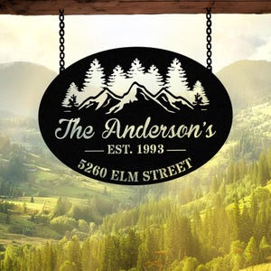 Peut inclure: Panneau ovale en métal noir avec un motif de montagne et de pins, suspendu par des chaînes. Le panneau indique "The Anderson's EST. 1993 5260 ELM STREET" dans une police décorative. Le fond montre un paysage de montagne.