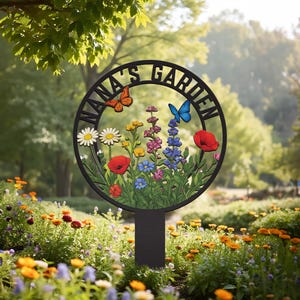 May include: A black metal garden sign with the words "NANA'S GARDEN" in a circular design. The sign features colorful flowers, two butterflies, and a stake for placement in a garden. The sign is surrounded by a garden of colorful flowers.