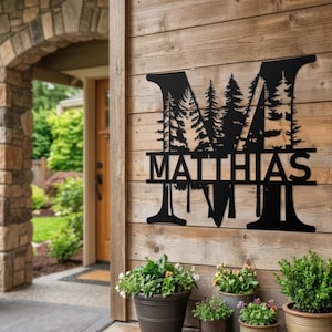 Puede incluir: Arte de pared de metal negro con el nombre "MATTHIAS" debajo de una letra "M" estilizada con una silueta de bosque. La obra de arte está montada en una pared de madera, con plantas en macetas delante.