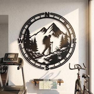 May include: A circular black metal wall art piece featuring a silhouette of a hiker with a backpack and trekking poles, set against a mountain and pine tree landscape. The outer ring displays cardinal directions N, S, E, W, NE, NW, SE, and SW.