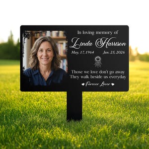 以下が含まれることがあります： 女性の写真とテキストが書かれた黒い記念ガーデンステーク。「In loving memory of Linda Harrison」というテキストと日付、引用句「Those we love don't go away. They walk beside us everyday. Forever Lover.」が書かれています。