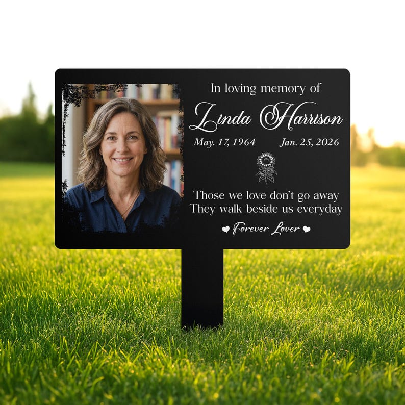 以下が含まれることがあります： 女性の写真とテキストが書かれた黒い記念ガーデンステーク。「In loving memory of Linda Harrison」というテキストと日付、引用句「Those we love don't go away. They walk beside us everyday. Forever Lover.」が書かれています。