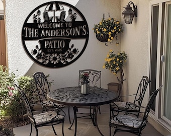 May include: Black metal patio sign with the text "WELCOME TO THE ANDERSON'S PATIO EST. 2025". The sign features a circular design with an umbrella, chairs, and floral accents. A patio set with a table and chairs is visible.