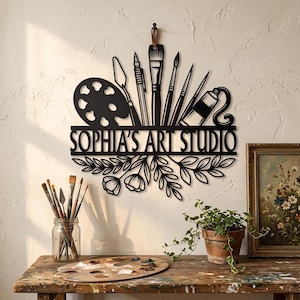 以下が含まれることがあります： SOPHIA'S ART STUDIOという文字が刻まれた黒い金属製の壁掛け看板。上部には絵の具パレット、筆、パレットナイフ、絵の具チューブのシルエットが施され、下部には葉と花の装飾的なフローラルデザインがあしらわれています。