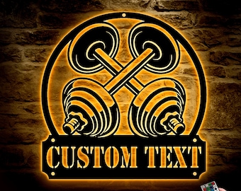May include: A black metal wall sign with a circular design featuring two crossed dumbbells. The sign has a custom text area that says "CUSTOM TEXT". The sign is illuminated with warm white light.