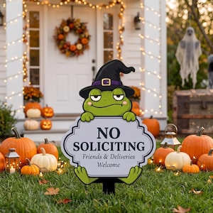 May include: A green frog yard sign wearing a black witch hat, holding a white sign with a decorative border that reads NO SOLICITING Friends & Deliveries Welcome in black text. The sign is positioned on a stake in a grassy lawn area.