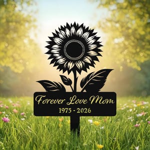 May include: Black metal garden stake featuring a sunflower design, with the inscription "Forever Love Mom" and the years 1975-2026. The stake is placed in a grassy area with a blurred background of trees.